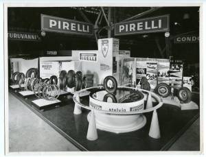 View of the Pirelli stand at the XXIV Geneva Motor Show in 1954. The image shows the Stelvio tyre on a revolving installation attached to a mirror base. In the background are signs with indications and photographs of the last two victories at the world auto championships and 4 Pirelli tyres fitted to the Ferrari of Umberto Maglioli during the Carrera Messicana. To the left, a display of the complete Pirelli tyre range.<br />