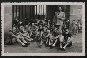 Ritratto di gruppo di corridori, seduti per terra: in piedi, dietro di loro, vi è un uomo, probabilmente un dirigente sportivo o un ufficiale di gara