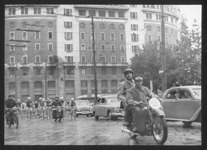 Il passaggio dei corridori e delle moto che precedono la corsa a Milano: è visibile il palazzo all'angolo tra viale Abruzzi e piazzale Loreto che ospita, tra il 1945 e il 1960, la sede centrale della Pirelli