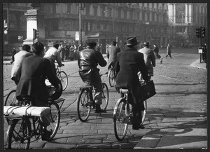 Il passaggio di alcuni uomini in bicicletta in piazza Cordusio: sulla sinistra è parzialmente visibile il monumento a Giuseppe Parini, sullo sfondo è visibile il Palazzo della Banca d'Italia