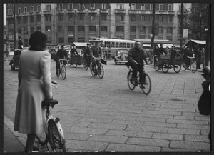 Il passaggio di biciclette, furgoncini a pedali e veicoli a motore in piazzale Loreto: sullo sfondo è visibile il palazzo all'angolo tra piazzale Loreto e viale Abruzzi, che ospita tra il 1945 e il 1960 la sede centrale della Pirelli
