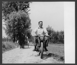 Un uomo in sella a una motocicletta in una strada di campagna, seguito da un carro trainato a cavallo