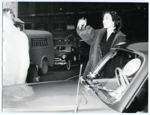 L'attrice Maria Fiore accanto a un'auto durante il passaggio da Roma delle vetture partecipanti al Rally di Monte Carlo del 1954. Sullo sfondo, un furgone con logo Pirelli.