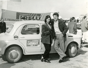 Gli attori Emma Danieli e Roberto Risso accanto alla vettura 06 del Rally di Monte Carlo 1954, Scuderia Titanus - Equipe 1. Sullo sfondo, uno striscione pubblicitario con logo Pirelli.