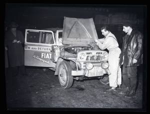 Un meccanico versa del liquido nel vano motore della Fiat Campagnola targata 127012.TO durante il III Rally del Sestriere del 22-25 febbraio 1952. Un uomo lo osserva.
